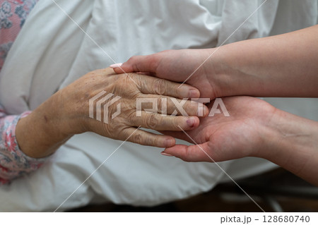 A young womans hands gently hold the hands of an elderly female patient in a hospital bed, symbolizing care, support, and compassion. 128680740