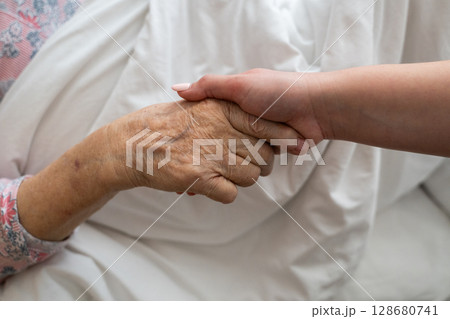A young womans hands gently hold the hands of an elderly female patient in a hospital bed, symbolizing care, support, and compassion. 128680741