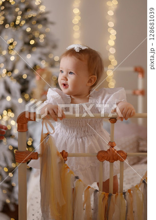 Caucasian baby girl in white dress sitting on bed near Christmas tree in cozy bedroom Caucasian baby girl in white dress sitting on bed near Christmas tree in cozy bedroom 128681039