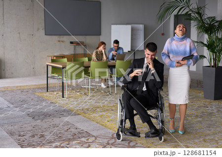 In a professional office setting, a businesswoman helping her director, who is sitting in a wheelchair and using a tablet, while their colleagues collaborate in the background. In a professional office setting, a businesswoman helping her director, who is sitting in a wheelchair and using a tablet, while their colleagues collaborate in the background. 128681154