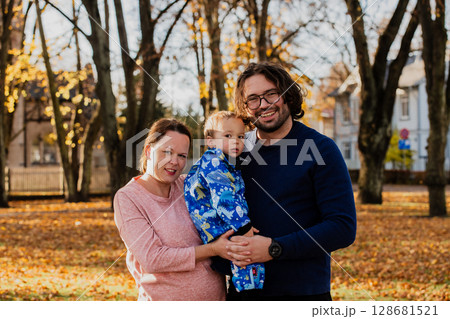 On a sunny autumn day in the park, a mother and father lovingly embrace their young son, sharing moments of laughter and joy as they play together 128681521