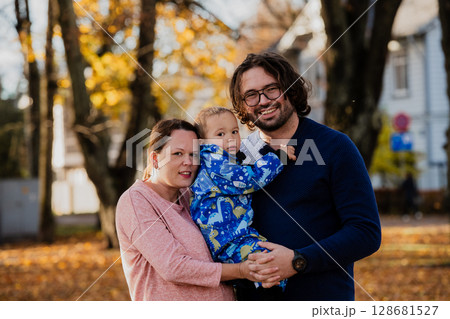 On a sunny autumn day in the park, a mother and father lovingly embrace their young son, sharing moments of laughter and joy as they play together 128681527