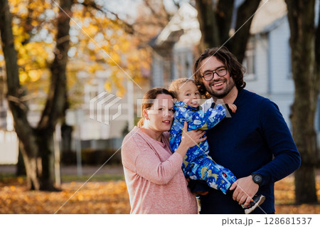 On a sunny autumn day in the park, a mother and father lovingly embrace their young son, sharing moments of laughter and joy as they play together 128681537