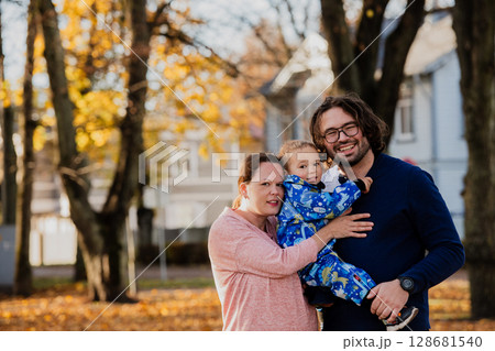 On a sunny autumn day in the park, a mother and father lovingly embrace their young son, sharing moments of laughter and joy as they play together 128681540