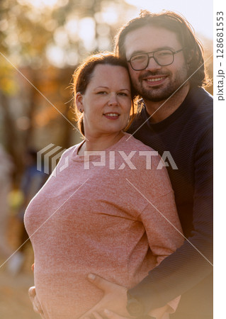 A man lovingly embraces his pregnant wife in the park, sharing a peaceful moment of warmth and affection  128681553
