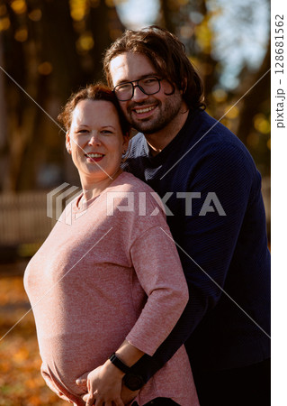 A man lovingly embraces his pregnant wife in the park, sharing a peaceful moment of warmth and affection  128681562