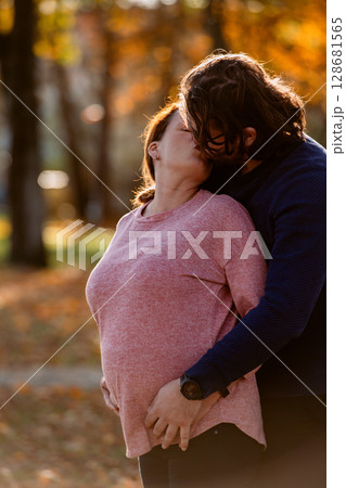A man lovingly embraces his pregnant wife in the park, sharing a peaceful moment of warmth and affection  128681565