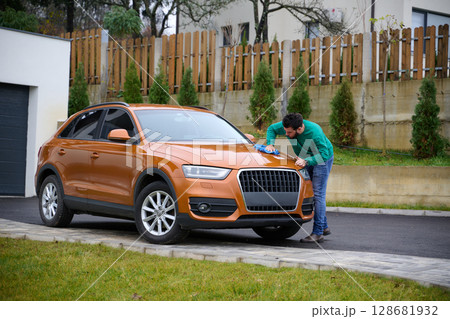 A man meticulously washes and cleans his car in the driveway of his home, showcasing pride in his vehicle and outdoor space. 128681932