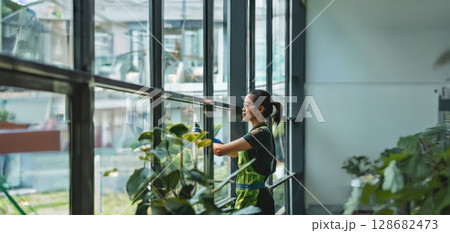 Professional Window Cleaner in Protective Gear Cleaning Interior Windows with Plants in the Background.Generated image 128682473