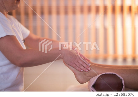 Foot Massage Therapy in a Relaxing Spa Setting. A professional massage therapist in a white uniform provides a soothing foot massage to a client in a calm and serene spa environment 128683076