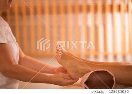 Foot Massage Therapy in a Relaxing Spa Setting. A professional massage therapist in a white uniform provides a soothing foot massage to a client in a calm and serene spa environment 128683080
