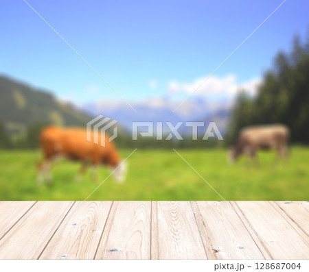 Old wooden table and cows grazing in a mountain meadow. Empty wooden table top. Rustic wooden board and View of mountain scenery in Alps on sunny day. Product display template. Copy space for text 128687004