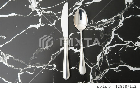 Luxury silver knife and spoon on black marble background, elegant modern cutlery set for fine dining restaurant or exclusive event, top view with copy space 128687112