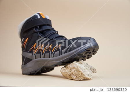 Black trekking shoe standing on small stone. Hiking boot on beige background. Outdoor footwear concept Black trekking shoe standing on small stone. Hiking boot on beige background. Outdoor footwear concept 128690320