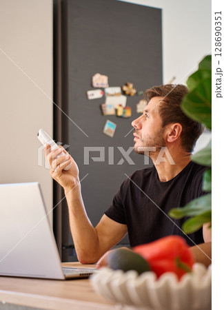 Man working on laptop at home and using remote control to adjust temperature on air conditioner. Person struggling with heat while working at home office 128690351
