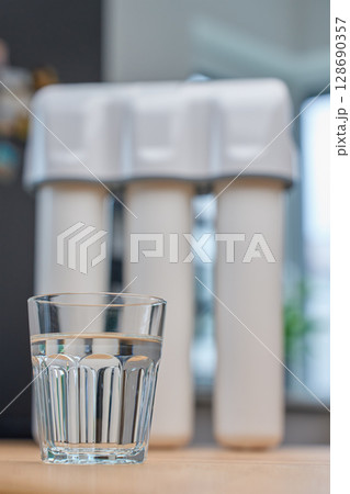 Modern water filtration system with three cartridges and glass of clean water on kitchen table. Home water purification, healthy lifestyle and drinking water safety concept 128690357