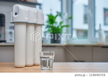 Modern water filtration system with three cartridges and glass of clean water on kitchen table. Home water purification, healthy lifestyle and drinking water safety concept 128690358