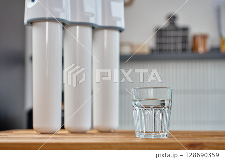 Modern water filtration system with three cartridges and glass of clean water on kitchen table. Home water purification, healthy lifestyle and drinking water safety concept Modern water filtration system with three cartridges and glass of clean water on kitchen table. Home water purification, healthy lifestyle and drinking water safety concept 128690359