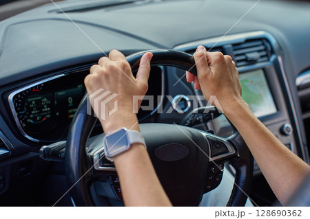 Close-up of hands holding steering wheel inside modern car with dashboard in background. Woman driving car 128690362