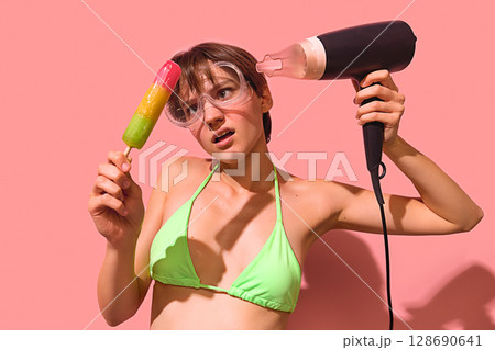 Confused woman melting rainbow popsicle with hairdryer wearing green bikini and goggles Confused woman melting rainbow popsicle with hairdryer wearing green bikini and goggles 128690641