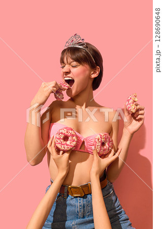 Laughing woman in tiara biting pink donut while wearing edible donut bra on pink background Laughing woman in tiara biting pink donut while wearing edible donut bra on pink background 128690648