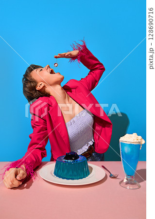 Woman in pink jacket joyfully reaching for cherry above blue jelly dessert and creamy cocktail Woman in pink jacket joyfully reaching for cherry above blue jelly dessert and creamy cocktail 128690716