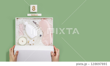 Womans day holiday flat lay with female hands hold white gift box with sleep care set. Self care package, craft gift box with mask for sleep, aroma candle and bubble bath. Flat lay, Copy space 128697891