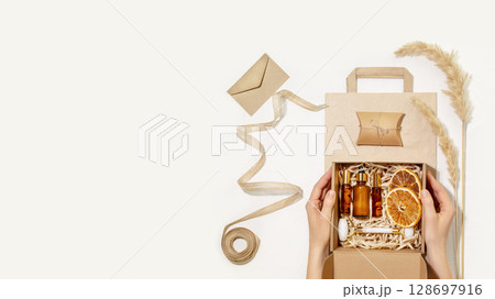 Female hands hold cardboard gift box with self care set with natural cosmetic on paper bag. Packaging goods from online store, delivery. Eco friendly packaging concept. Self care gift set. Copy space Female hands hold cardboard gift box with self care set with natural cosmetic on paper bag. Packaging goods from online store, delivery. Eco friendly packaging concept. Self care gift set. Copy space 128697916