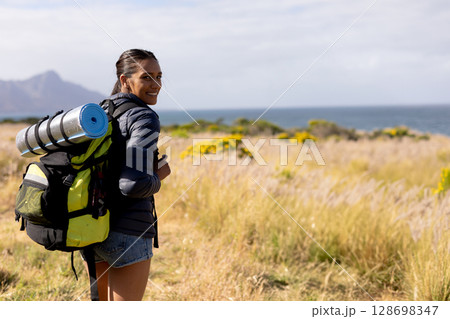 Smiling woman hiking with backpack and yoga mat in scenic nature, copy space 128698347