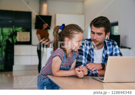 Work from home. Daughter interrupting her father while he is having a business online conversation on his laptop while sitting in the modern living room. 128698348