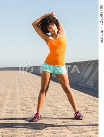 Woman stretching outdoors on sunny promenade, wearing bright athletic outfit 128698400
