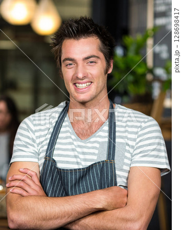 Barista smiling confidently in coffee shop, wearing striped apron, arms crossed Barista smiling confidently in coffee shop, wearing striped apron, arms crossed 128698417