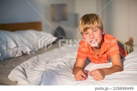 Smiling boy in orange shirt relaxing on bed at home, enjoying leisure time, copy space 128698449