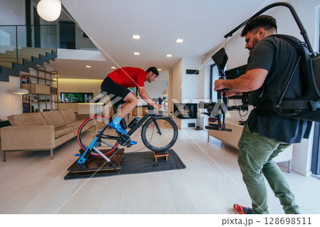 A cameraman filming an athlete riding a triathlon bike on a simulation machine in a modern living room. Training in pandemic conditions A cameraman filming an athlete riding a triathlon bike on a simulation machine in a modern living room. Training in pandemic conditions 128698511