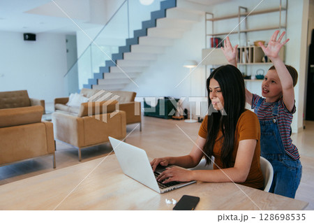 Mother with her daughter talking on laptop with family and friends while sitting in modern living room of big house. Mother with her daughter talking on laptop with family and friends while sitting in modern living room of big house. 128698535