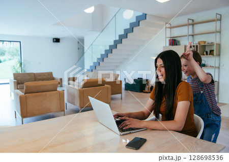 Mother with her daughter talking on laptop with family and friends while sitting in modern living room of big house. Mother with her daughter talking on laptop with family and friends while sitting in modern living room of big house. 128698536