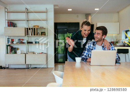A young married couple is talking to parents, family and friends on a video call via a laptop while sitting in the living room of their modern house. 128698580