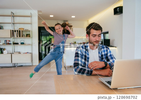 Work from home. Daughter interrupting her father while he is having a business online conversation on his laptop while sitting in the modern living room. Work from home. Daughter interrupting her father while he is having a business online conversation on his laptop while sitting in the modern living room. 128698702
