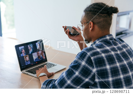 African American businessman in glasses sitting at a table in a modern living room, using a laptop and smartphone for business video chat, conversation with friends and entertainment. African American businessman in glasses sitting at a table in a modern living room, using a laptop and smartphone for business video chat, conversation with friends and entertainment. 128698711