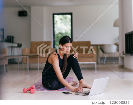 A woman in sportswear is sitting in the living room and preparing for online training while using a laptop. A woman in sportswear is sitting in the living room and preparing for online training while using a laptop. 128698825