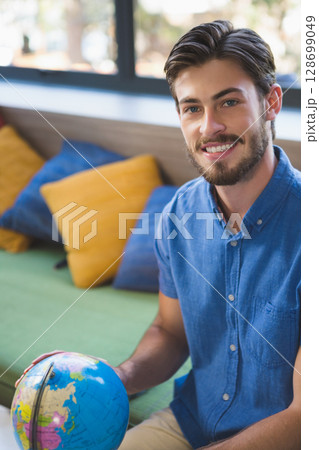 Smiling man sitting on green upholstered bench in modern lounge, holding small world globe 128699049