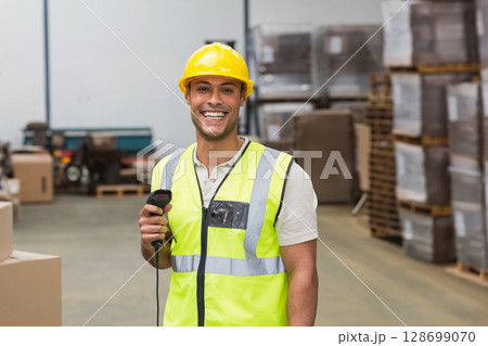 Smiling male warehouse worker holding barcode scanner in busy distribution center with pallets 128699070