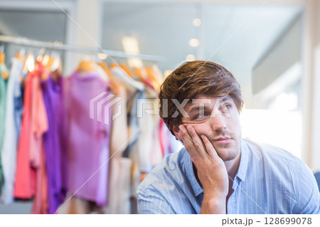 Leaning male shopper touching chin at clothing store, with pastel shirts on metal rack, copy space Leaning male shopper touching chin at clothing store, with pastel shirts on metal rack, copy space 128699078