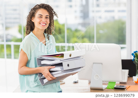 Smiling woman holding tall stack of binders and folders at modern office by window, copy space 128699079