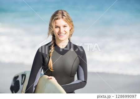 Smiling woman holding surfboard and wearing wetsuit on sandy beach, under clear blue sky 128699097