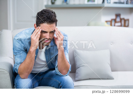Frowning man in twenties pressing temples on tufted sofa at home, with cushion, copy space 128699099