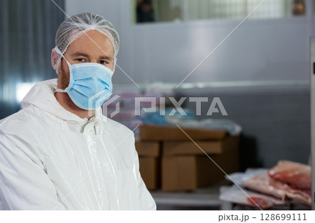 Standing man wearing protective suit and mask in processing plant, with meat, boxes, copy space Standing man wearing protective suit and mask in processing plant, with meat, boxes, copy space 128699111