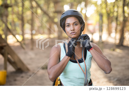Woman adjusting helmet strap in wooded ropes course, with protective gloves and safety harness 128699123