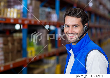 Smiling warehouse man coordinating inventory checks in aisles of boxes with headset, copy space 128699158