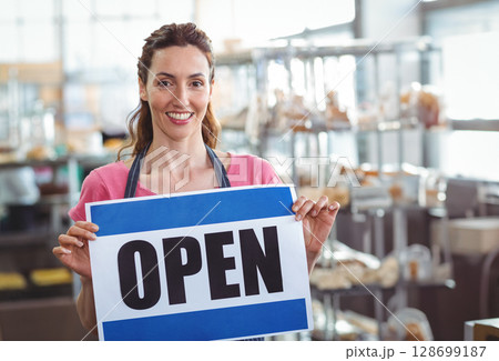Smiling woman baker holding OPEN sign inside bakery, with metal racks and pastries 128699187